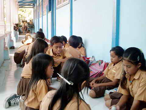 Kinder in gelb-brauner Uniform sitzen spielend unter dem Vordach der Schule.